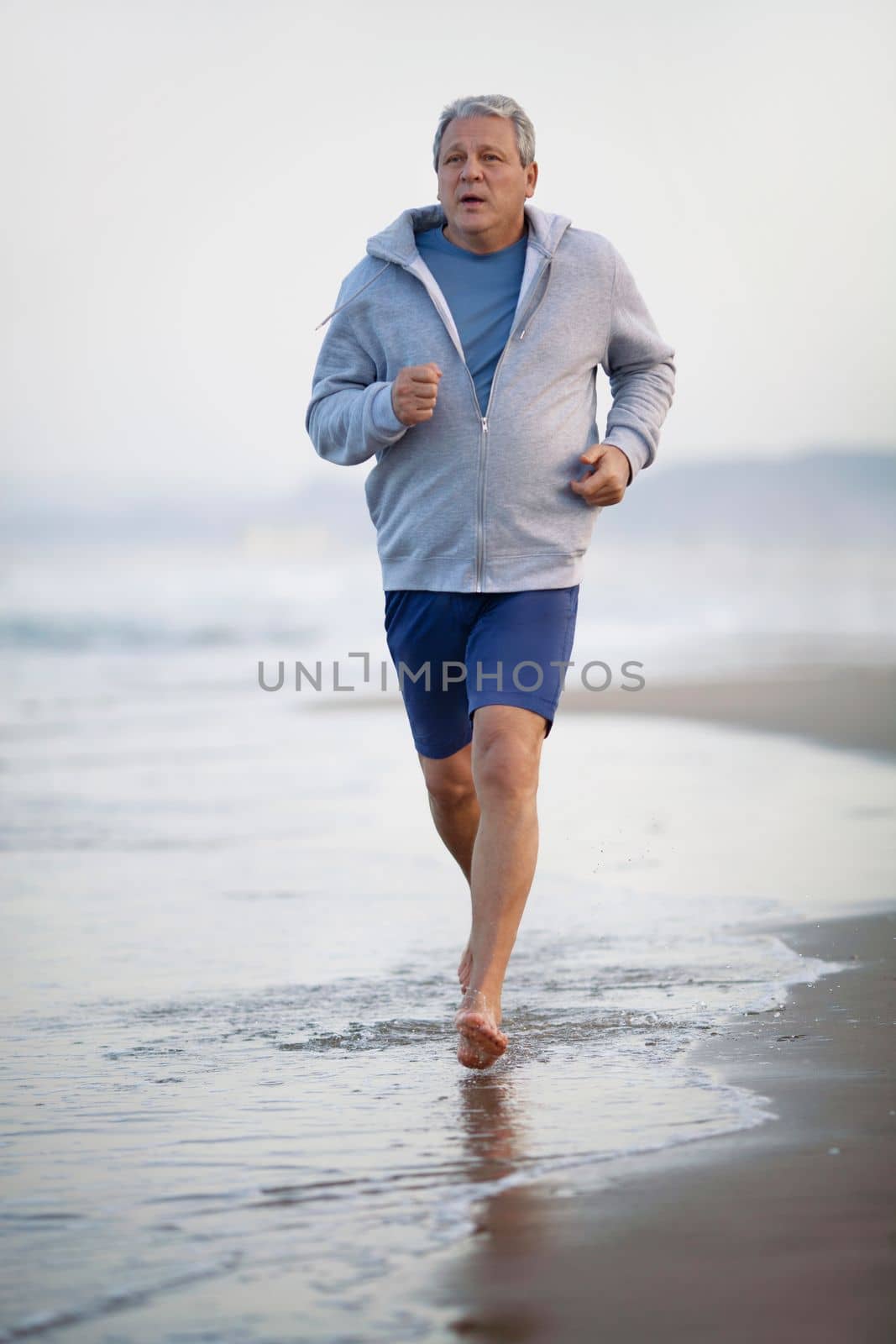 Barefoot senior man in shorts and hoodie jogging on the beach. Coastal sea waves washing his feet. Active and healthy lifestyle in retirement