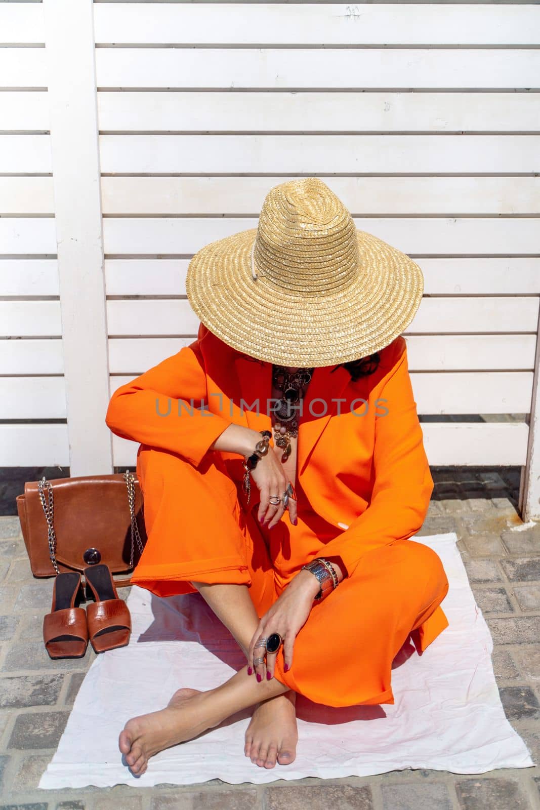 Stylish woman in an orange suit with a hat sits on a rug on a white striped background. On the hands are jewelry rings and bracelets, sandals and a bag stand side by side. by Matiunina