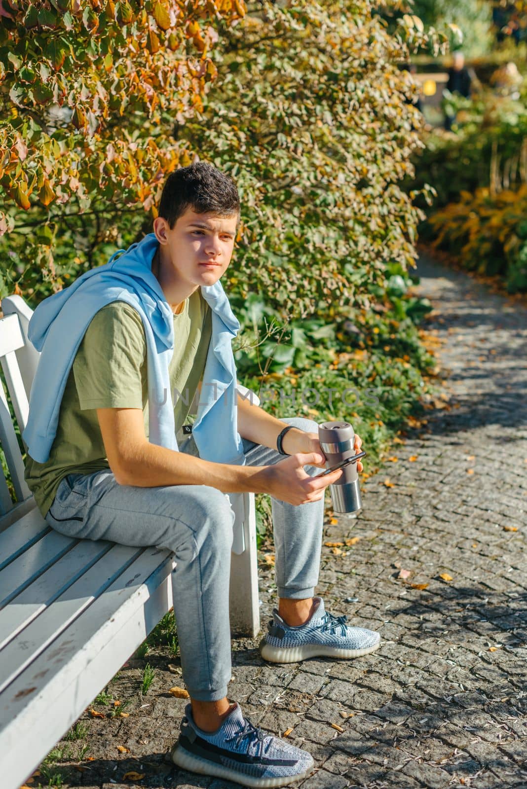 a teenager sits on a bench in the park drinks coffee from a thermo mug and looks into a phone. Portrait of handsome cheerful guy sitting on bench fresh air using device browsing media smm drinking latte urban outside outdoor.