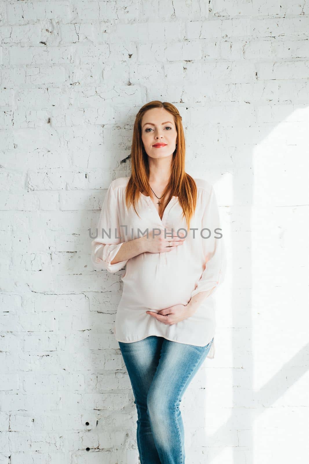 red-haired pregnant girl in a light blouse and blue jeans near the white wall of a large bright room