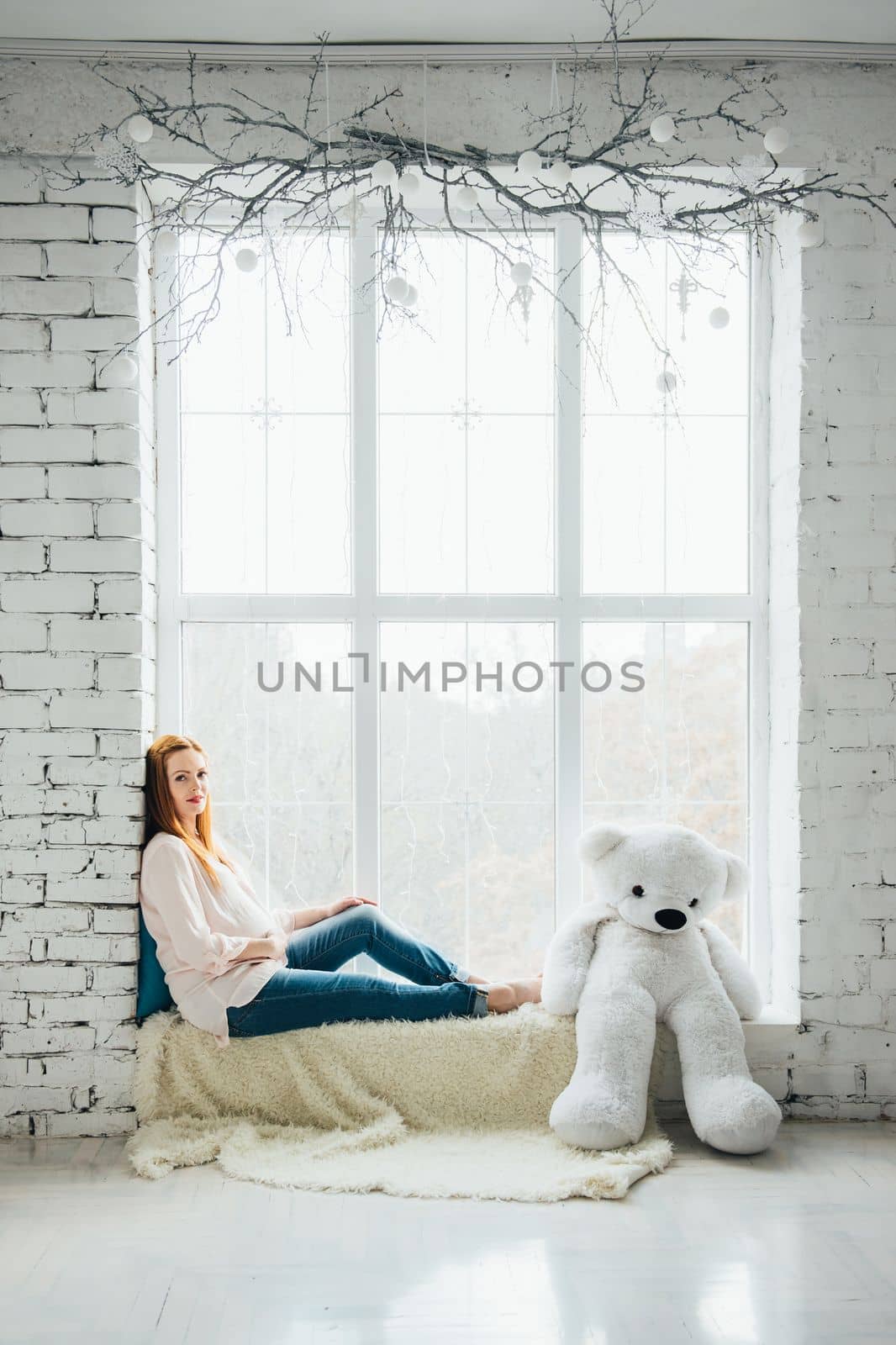 red-haired pregnant girl in a light blouse and blue jeans with a teddy bear on the window