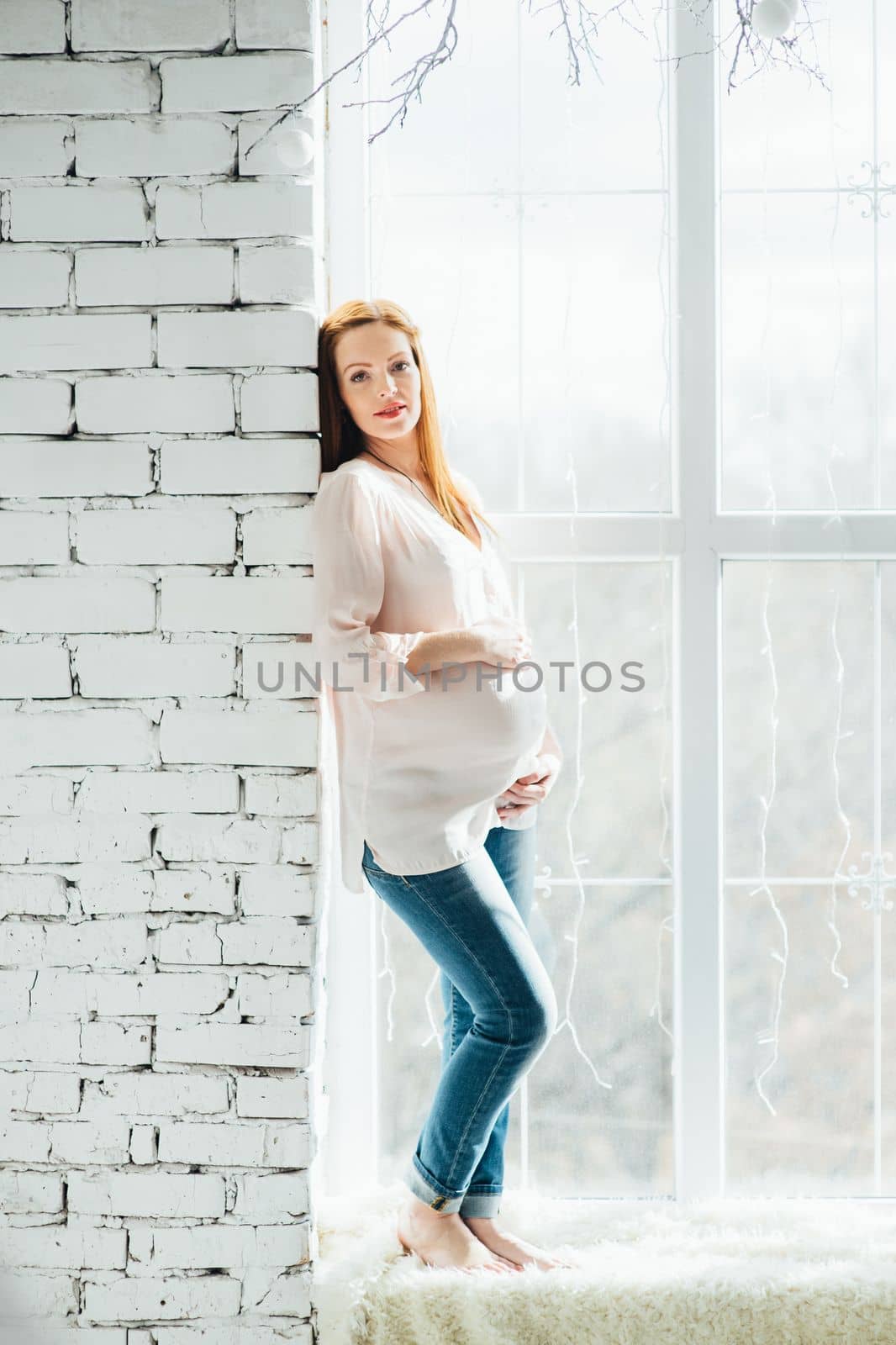 red-haired pregnant girl in a light blouse and blue jeans on a window near a brick wall
