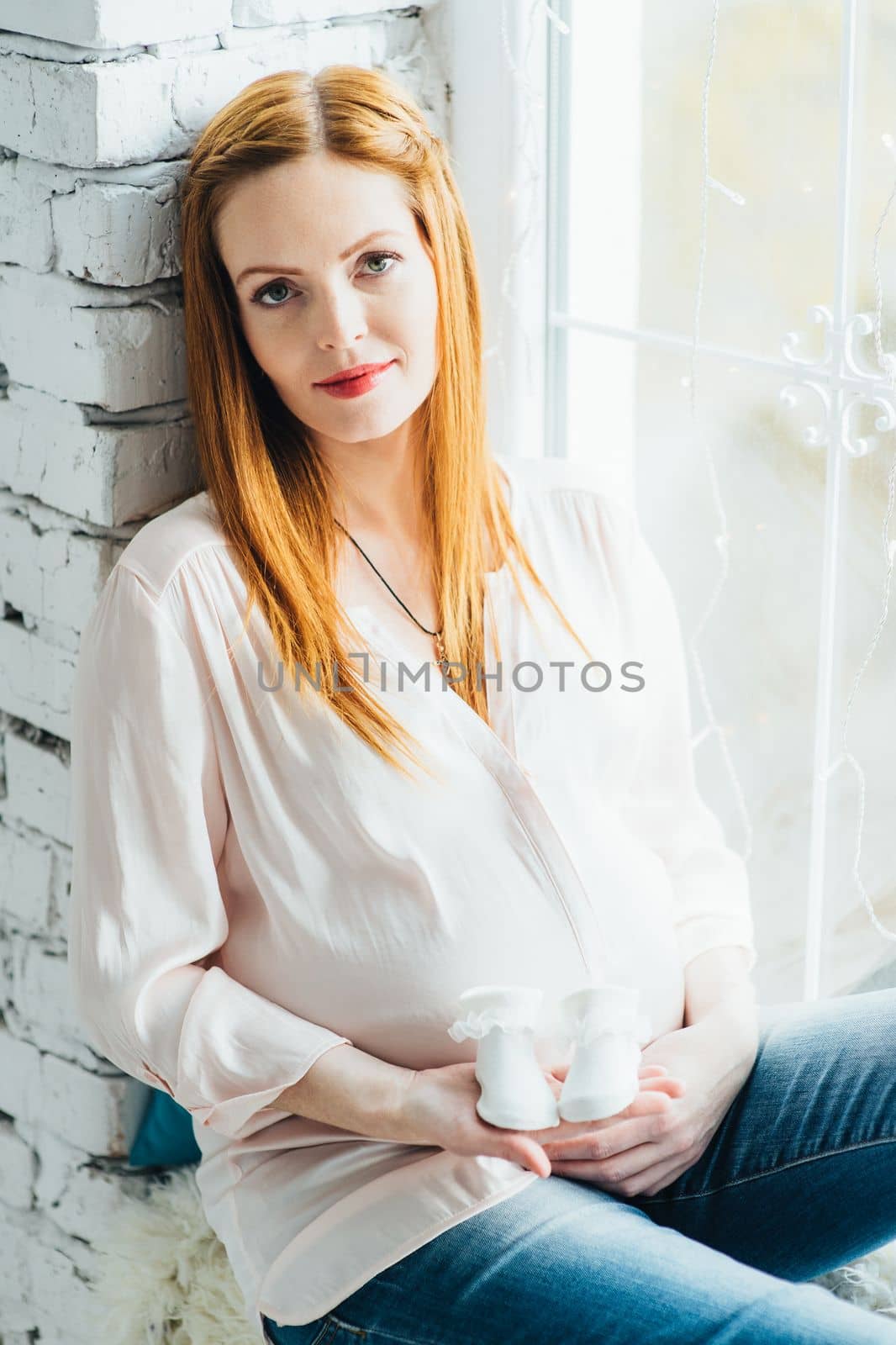 red-haired pregnant girl in a light blouse and blue jeans on a window near a brick wall