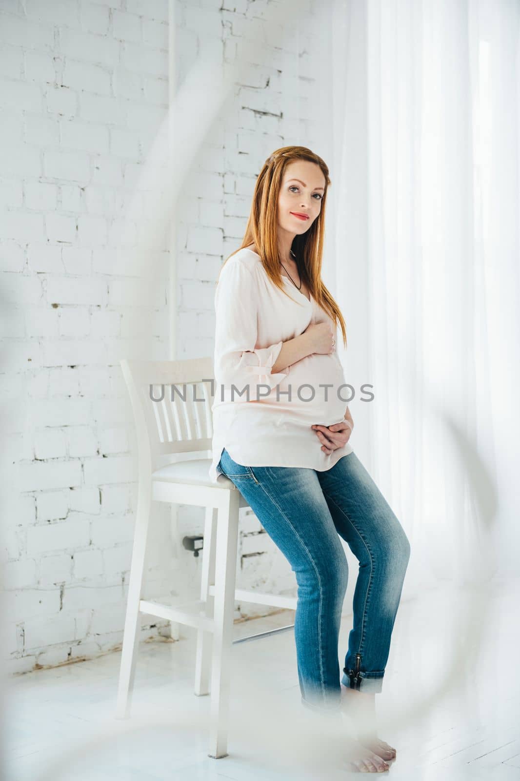 a red-haired pregnant girl in a light blouse and blue jeans sits on a high chair in a bright room
