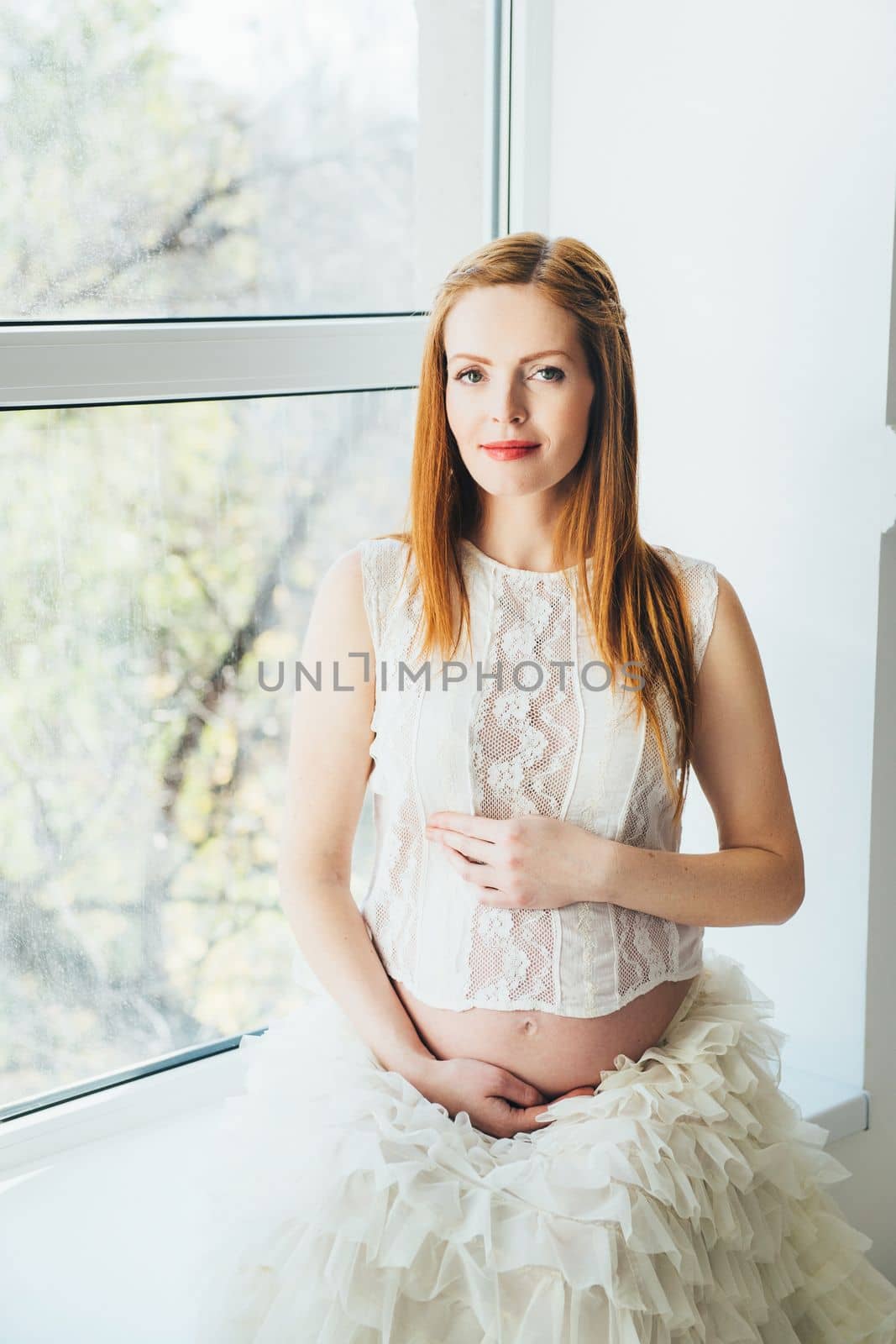 red-haired pregnant young girl in a white dress near the window of a bright room