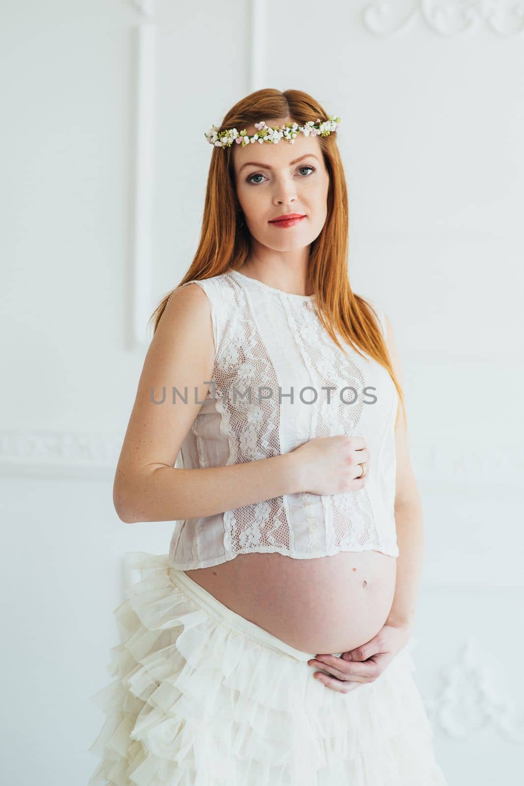 red-haired pregnant young girl in a white dress near the white wall of a large bright room