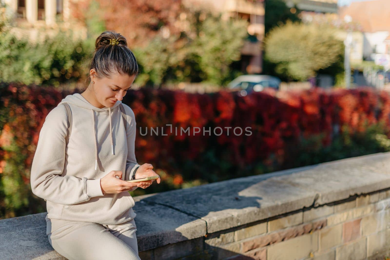 Young fashionable teenage girl with smartphone in park in autumn sitting at smiling. Trendy young woman in fall in park texting. Retouched, vibrant colors. Beautiful blonde teenage girl wearing casual modern autumn outfit sitting in park in autumn. Retouched, vibrant colors, brownish tones.