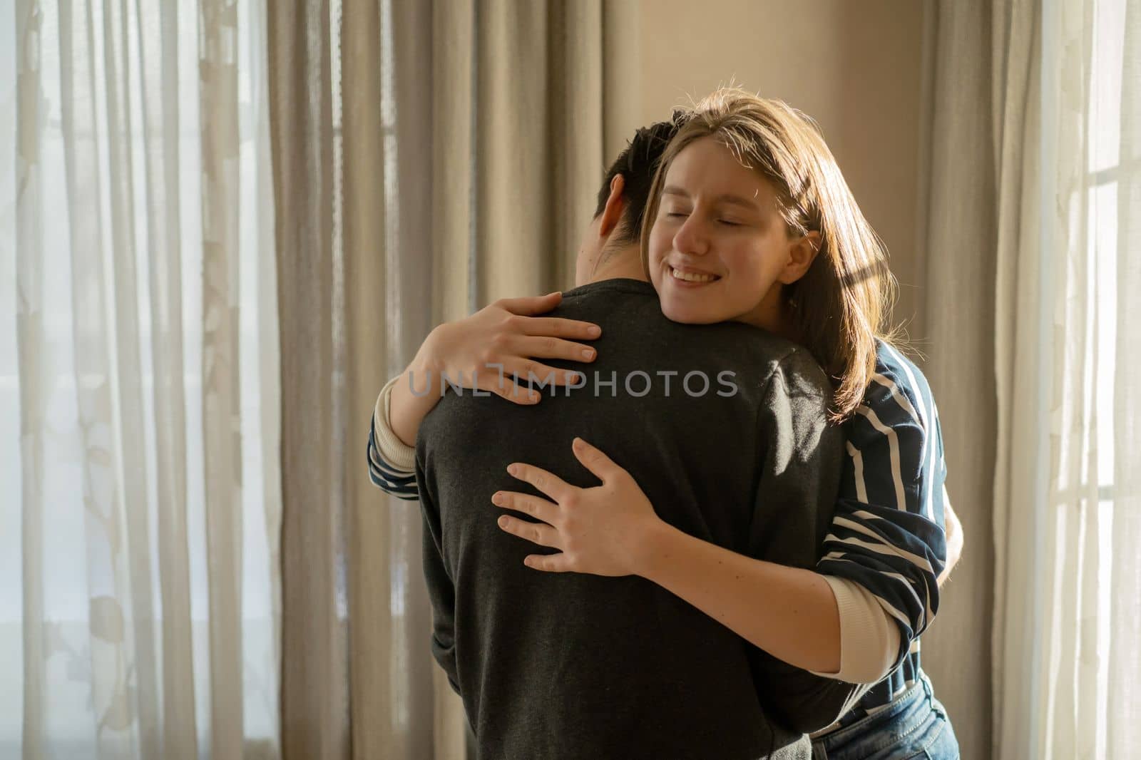 Happy young girl warmly hugs her boyfriend, rejoices at the meeting, stands near the window, smiley face in close-up, woman starts a romantic relationship. Husband and wife feel united, in a harmony.