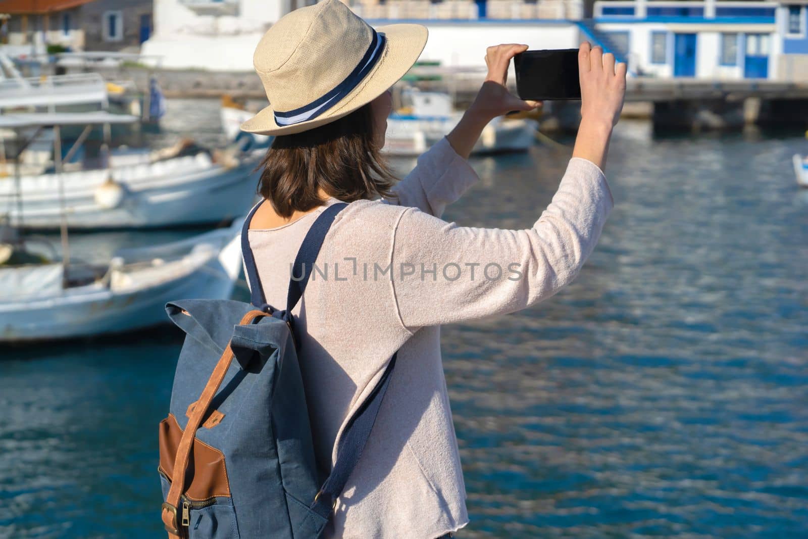 A young girl in a hat and with a backpack makes a photo of a beautiful bay with yachts and boats on the sea while traveling, a tourist walks along the shore next to the ocean and takes pictures.