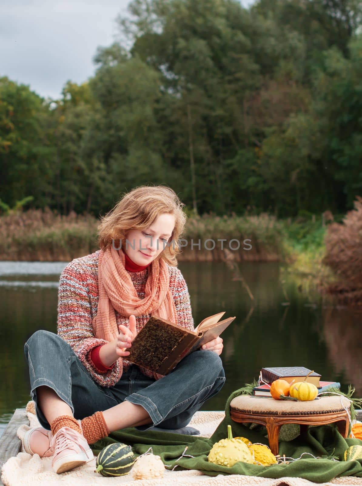 a beautiful red-haired girl reads a book on the embankment against the backdrop of autumn yellowing forests, fashion sweater. High quality photo