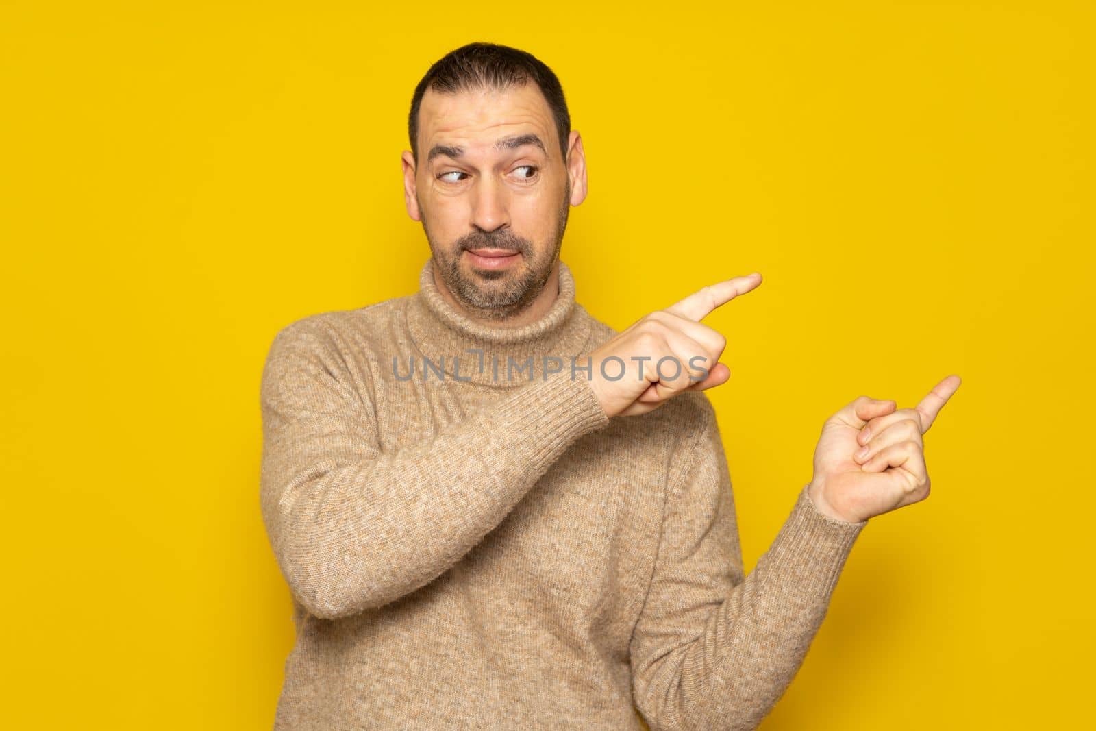 Hispanic man wearing a turtleneck sweater looking at the camera pointing with two hands and fingers to the side, isolated over yellow background.