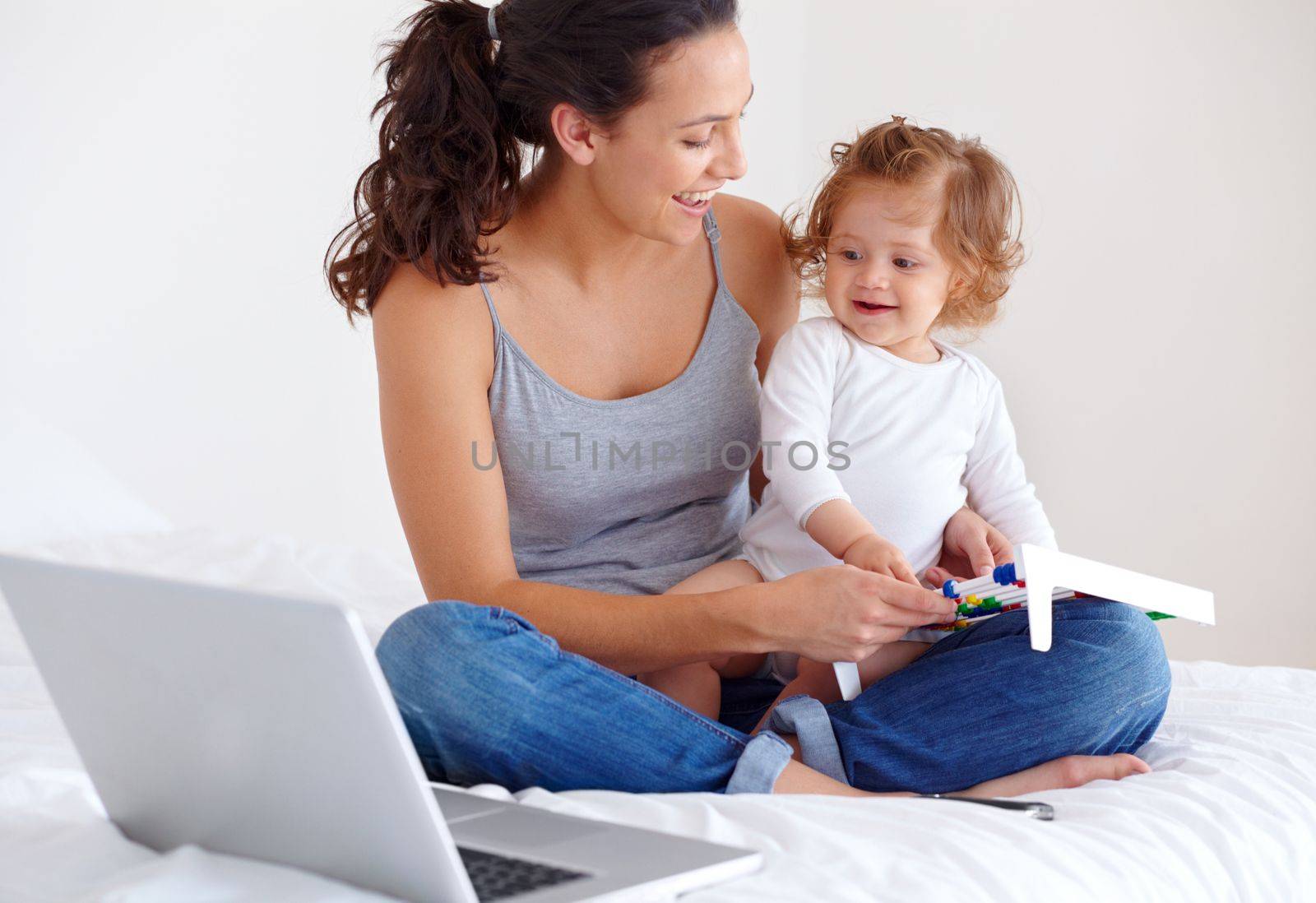 Juggling motherhood with work. A young mother sitting on a bed with her baby daughter on her lap and her laptop by her side