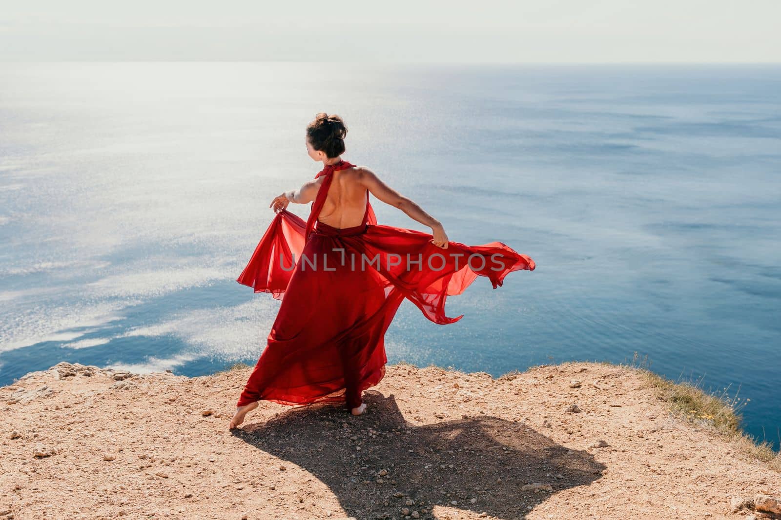 Side view a Young beautiful sensual woman in a red long dress posing on a rock high above the sea during sunrise. Girl on the nature on blue sky background. Fashion photo.