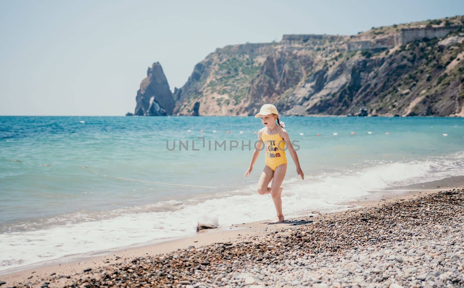 Cute little girl running along the seashore against a clear blue sea and rejoices in the rays of the summer sun. Beautiful girl in yellow swimsuit running and having fun on tropical beach