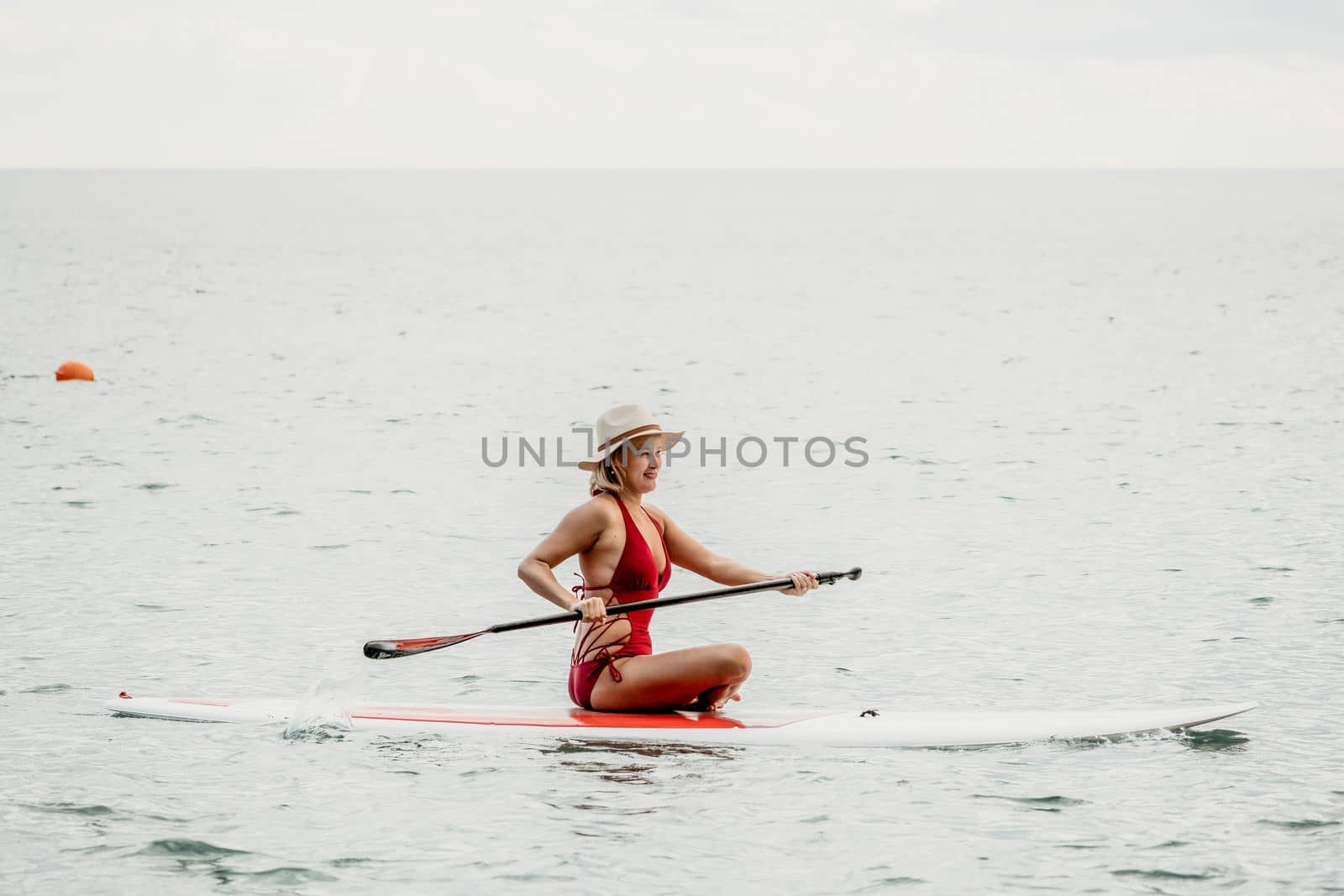 Woman in red bikini on sup board. Happy lady with blond hair in red bathing suit chilling and sunbathing by turquoise sea ocean on sunset. Holiday, vacation and recreational concept. by panophotograph