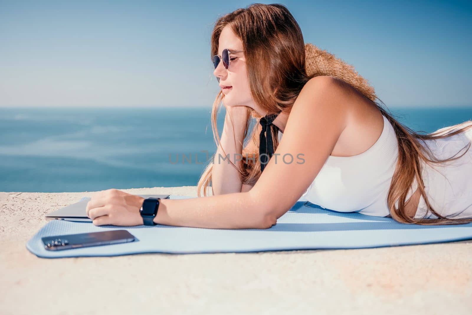 Successful business woman in yellow hat working on laptop by the sea. Pretty lady typing on computer at summer day outdoors. Freelance, travel and holidays concept.