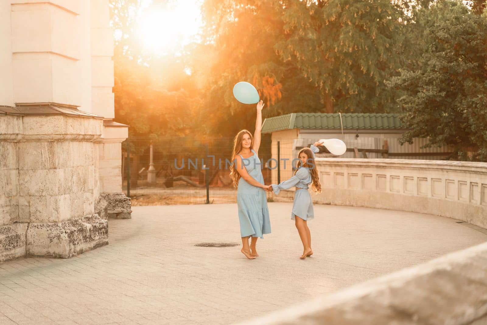 Daughter mother run holding hands. In blue dresses with flowing long hair, they hold balloons in their hands against the backdrop of a sunset and a white building