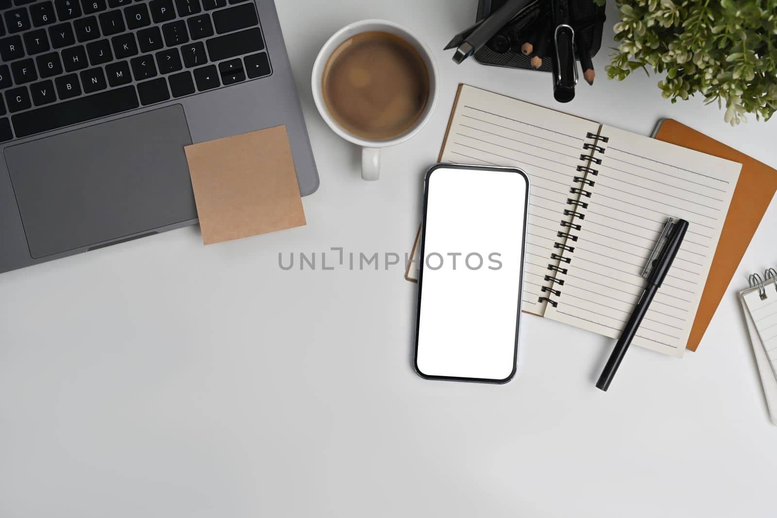 Flat lay, top view of smart phone with empty screen, notepad, laptop and coffee cup on white working desk by prathanchorruangsak