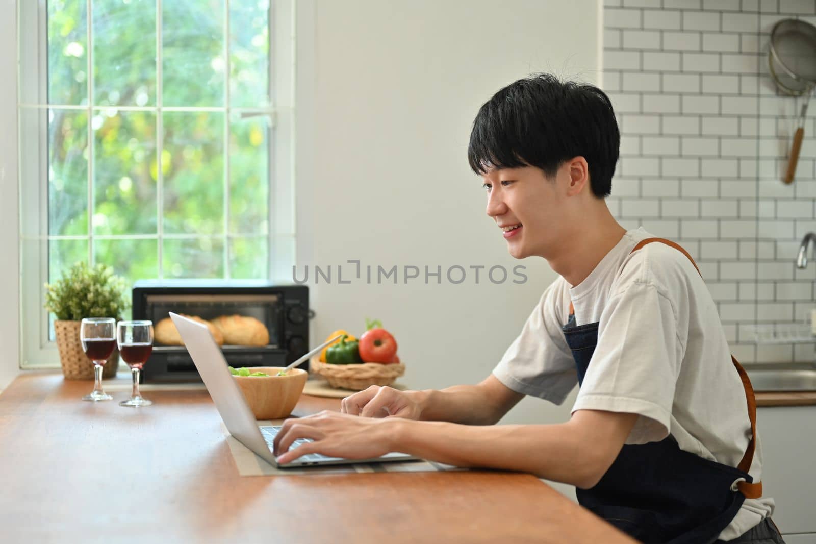 Side view of handsome man using laptop while sitting in modern sunny kitchen, Freelancer working remote from home.