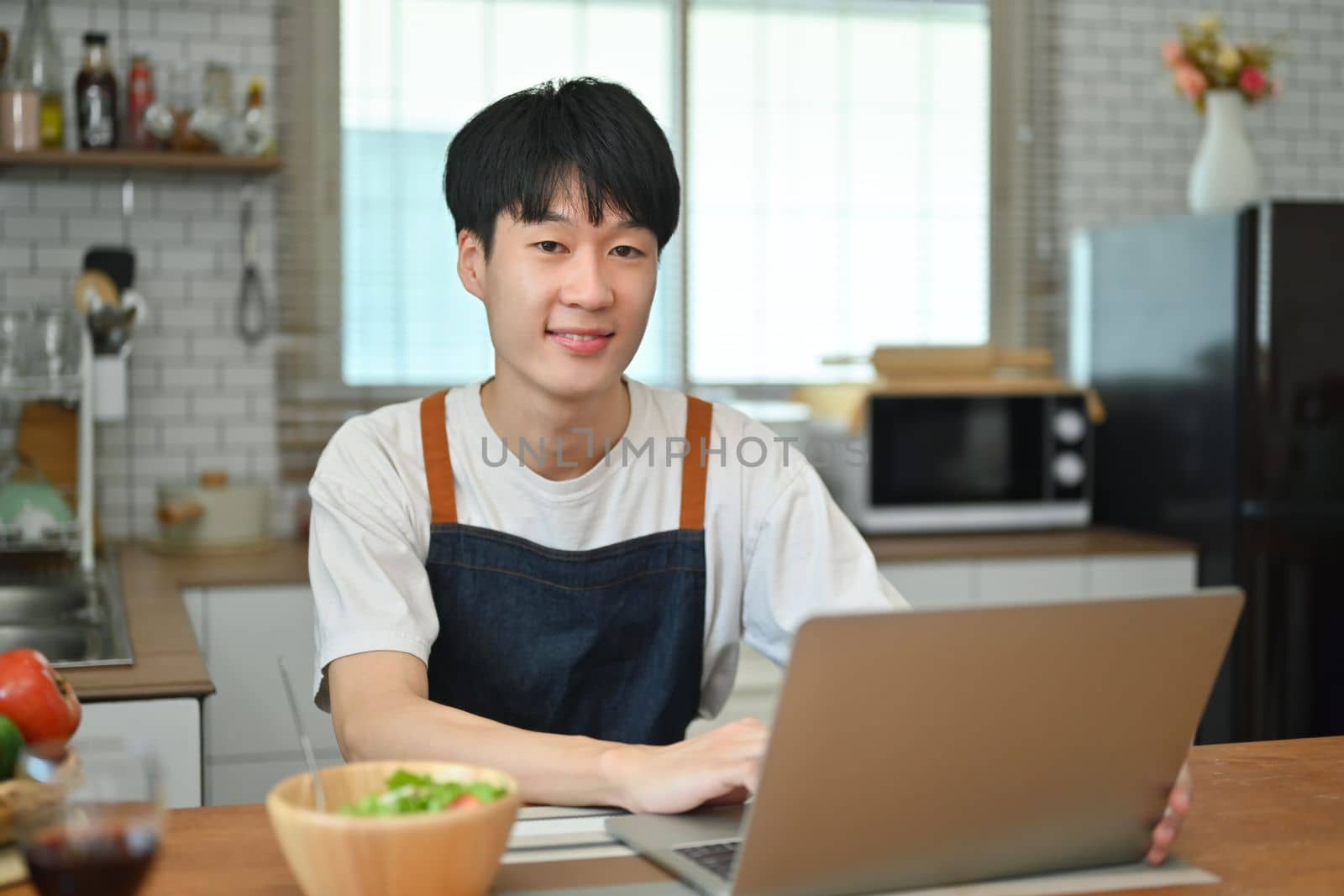 Asian man freelancer in apron and casual clothes working from home, sitting at kitchen worktop and using laptop.
