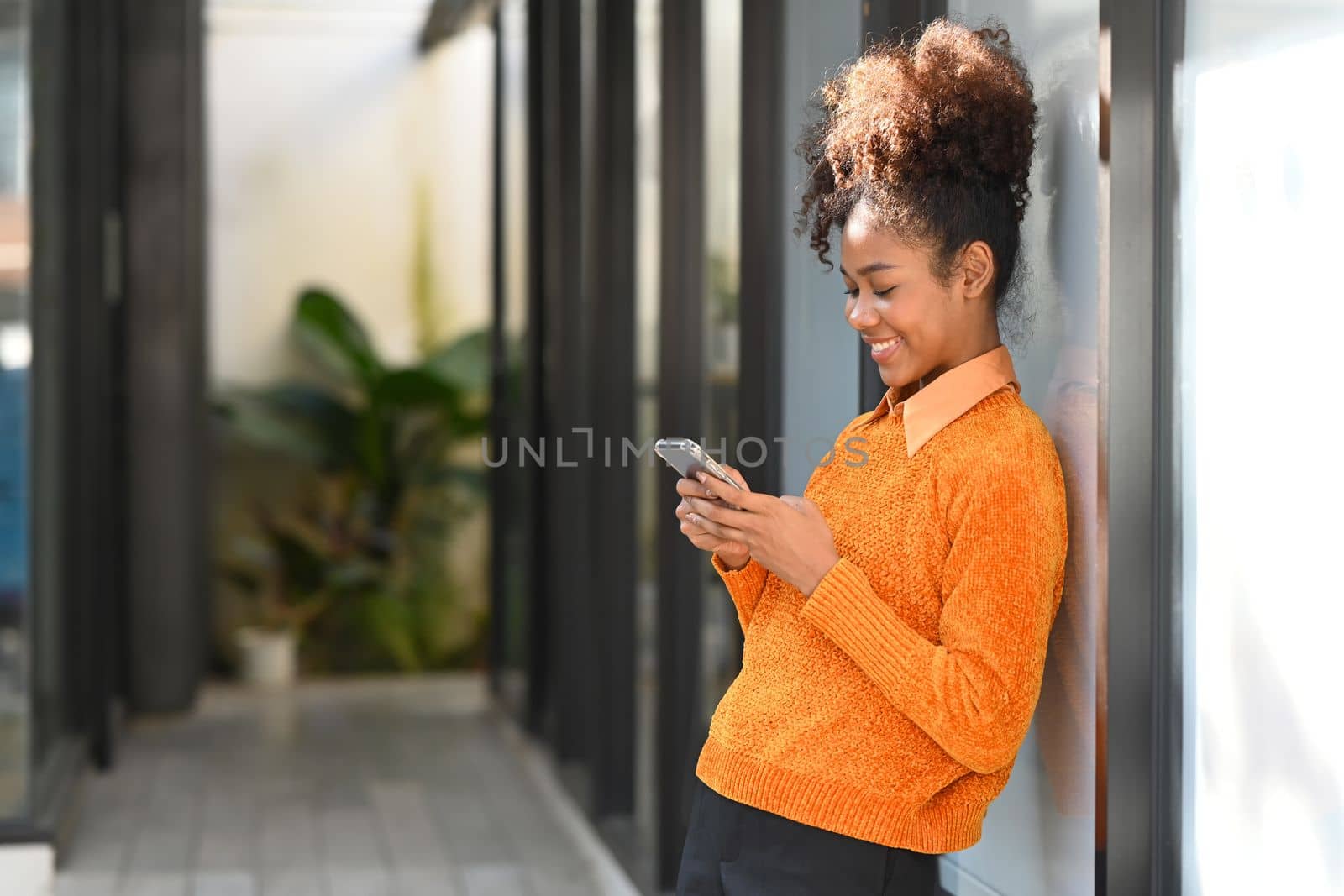 Image of smiling American African woman using mobile phone while standing outdoor near office building.