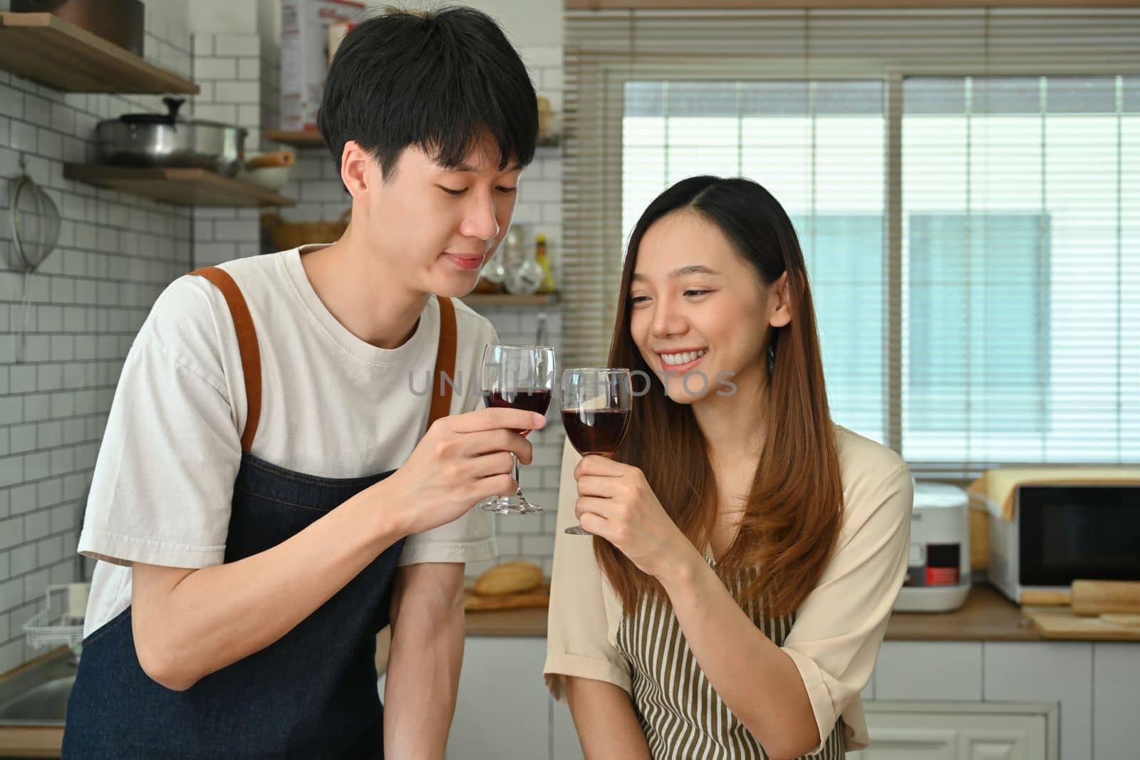 Romantic young couple clicking glasses of red wine ,laughing enjoying drinking red wine in modern kitchen by prathanchorruangsak