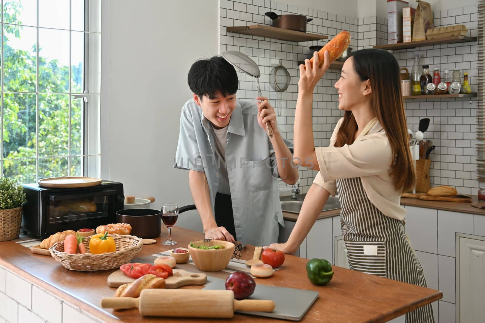 Playful young couple pretending fight with utensils tools while cooking in modern kitchen together.