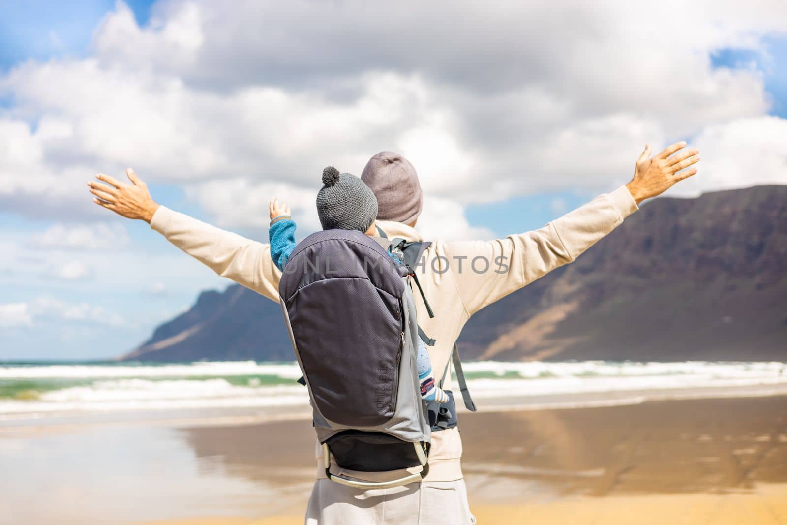 Young father rising hands to the sky while enjoying pure nature carrying his infant baby boy sun in backpack on windy sandy beach of Famara, Lanzarote island, Spain. Family travel concept