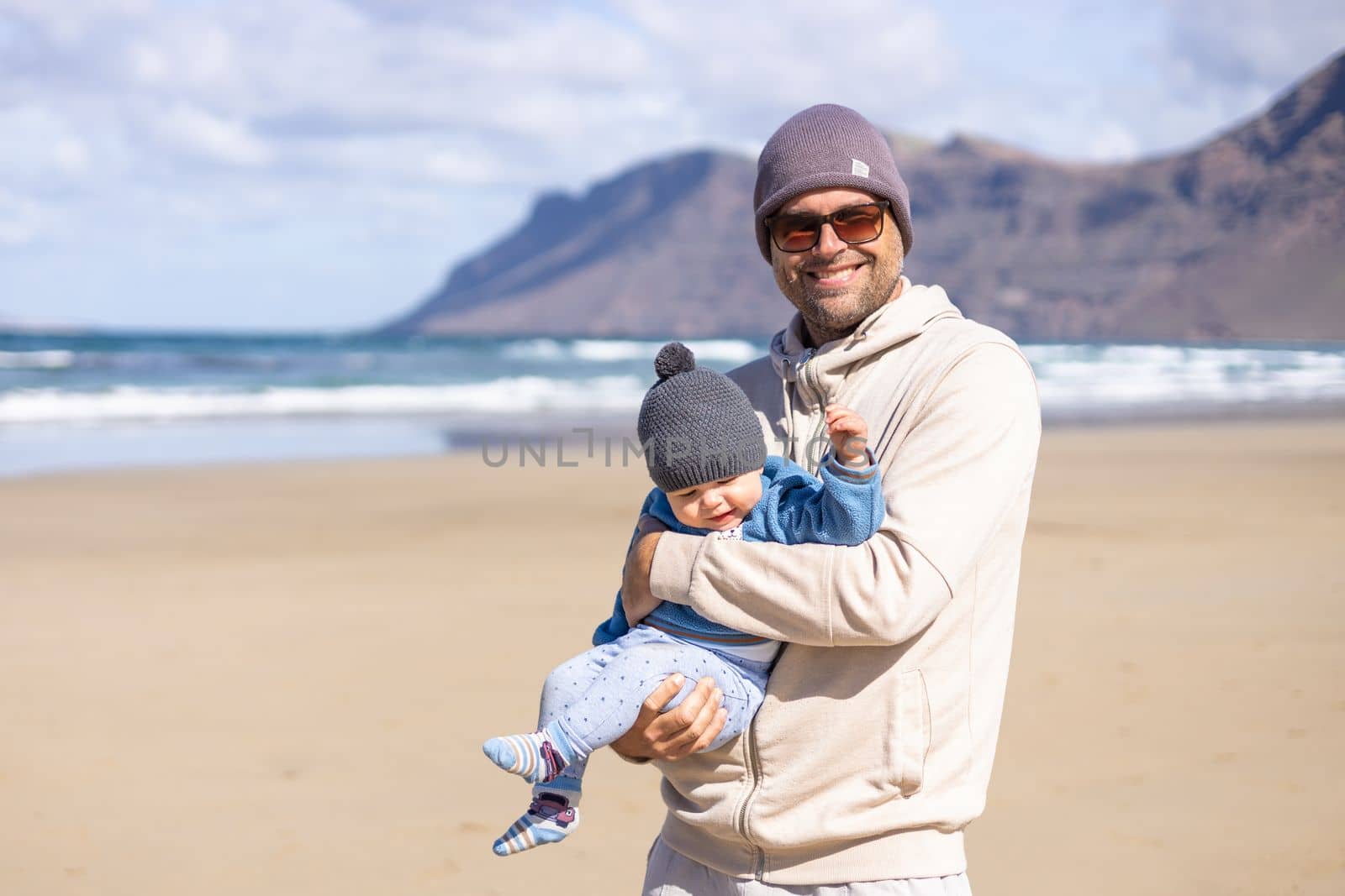 Father enjoying pure nature holding and playing with his infant baby boy sun in on windy sandy beach of Famara, Lanzarote island, Spain. Family travel and parenting concept