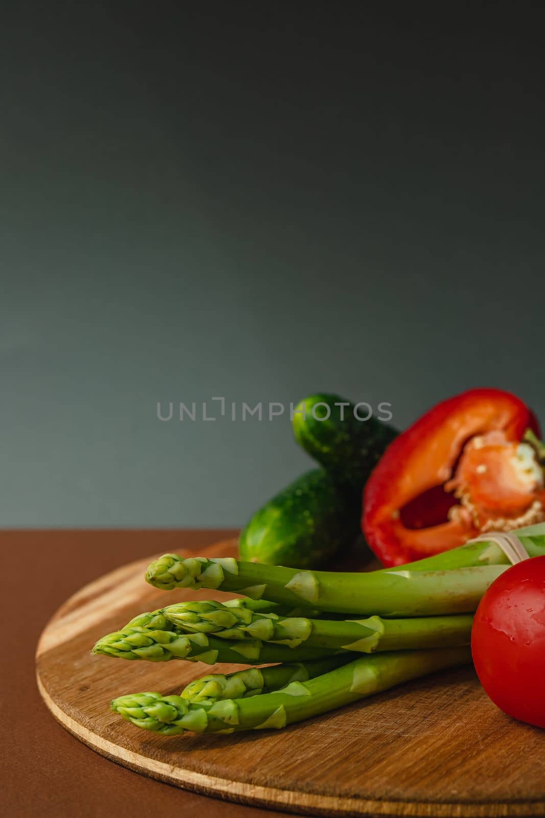 Vegetables lie on a wooden board: tomatoes, asparagus, cucumbers, red bell peppers. brown, dark gray background. place for text