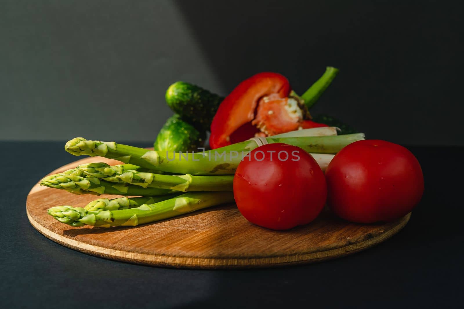 Vegetables lie on a wooden board: tomatoes, asparagus, cucumbers, red bell peppers. brown, dark gray background. place for text