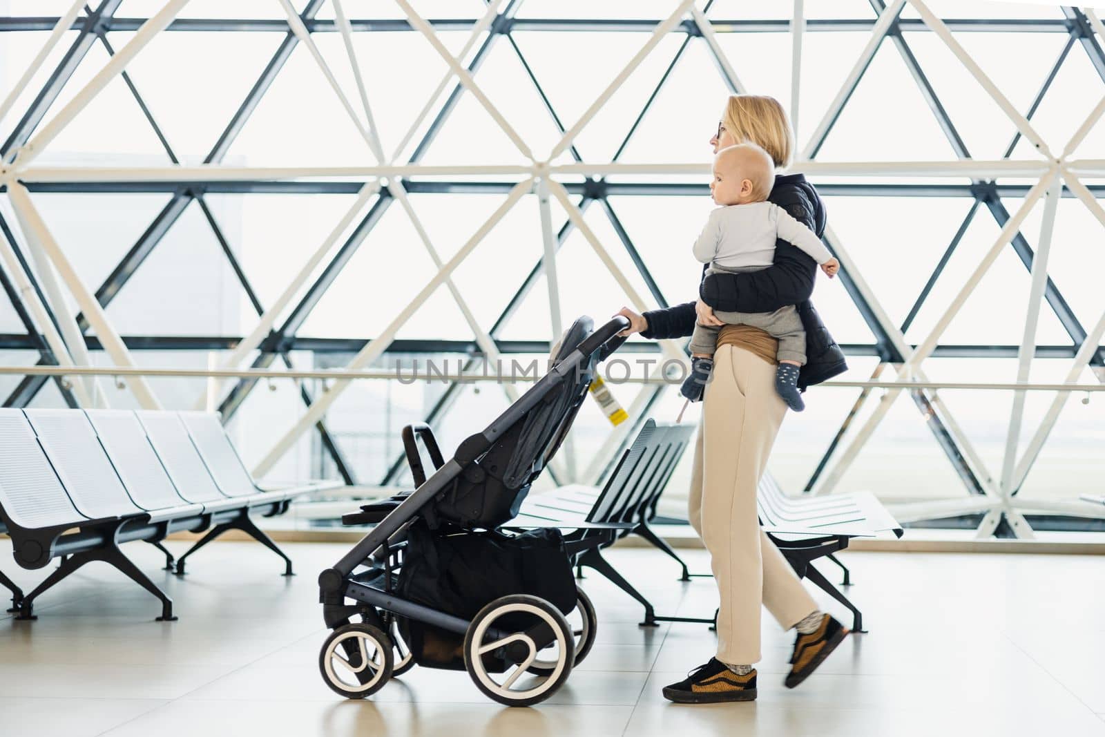 Mother carying his infant baby boy child, pushing stroller at airport departure terminal moving to boarding gates to board an airplane. Family travel with baby concept. by kasto