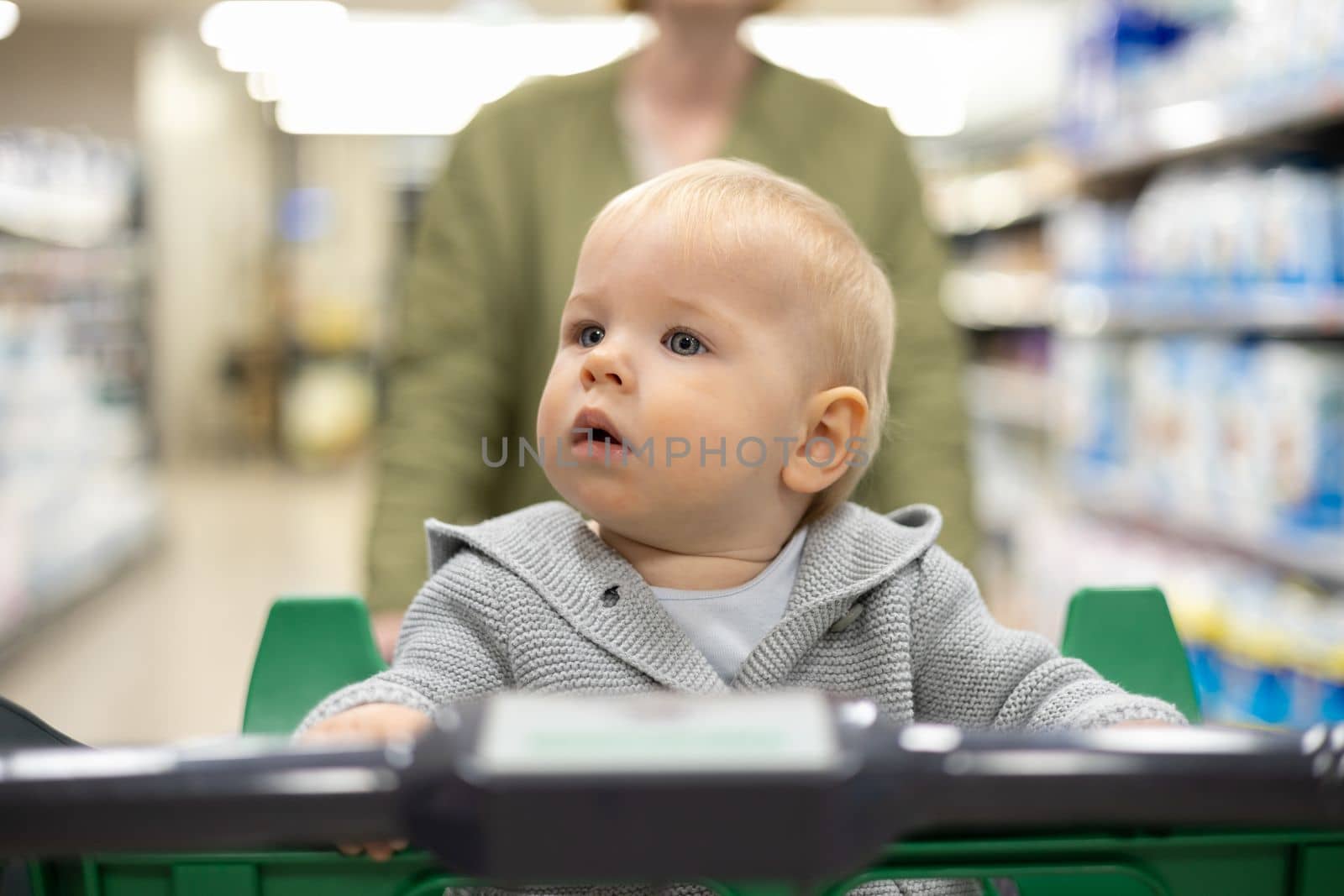 Mother pushing shopping cart with her infant baby boy child down department aisle in supermarket grocery store. Shopping with kids concept. by kasto