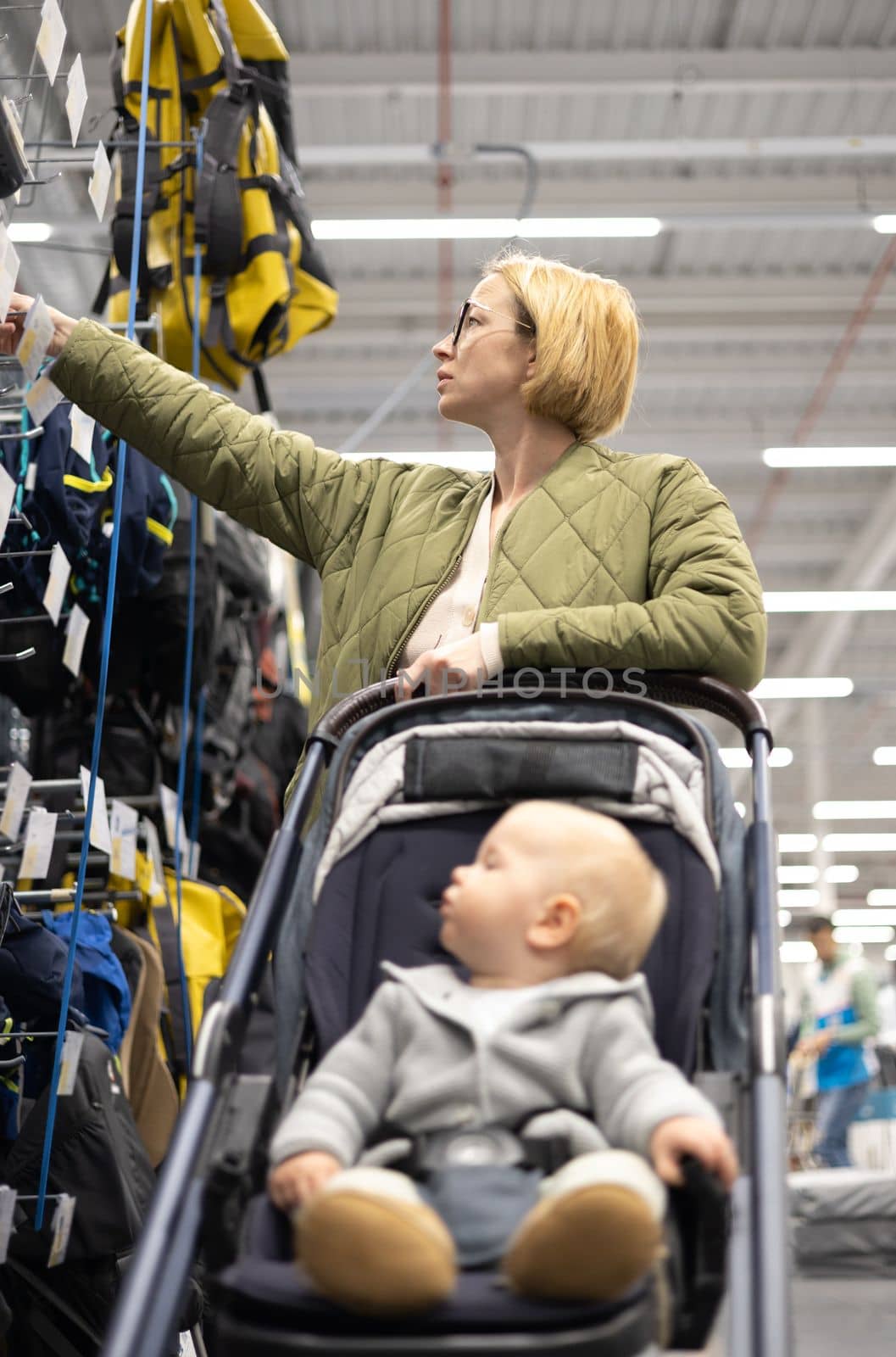 Casualy dressed mother choosing sporty shoes and clothes products in sports department of supermarket store with her infant baby boy child in stroller