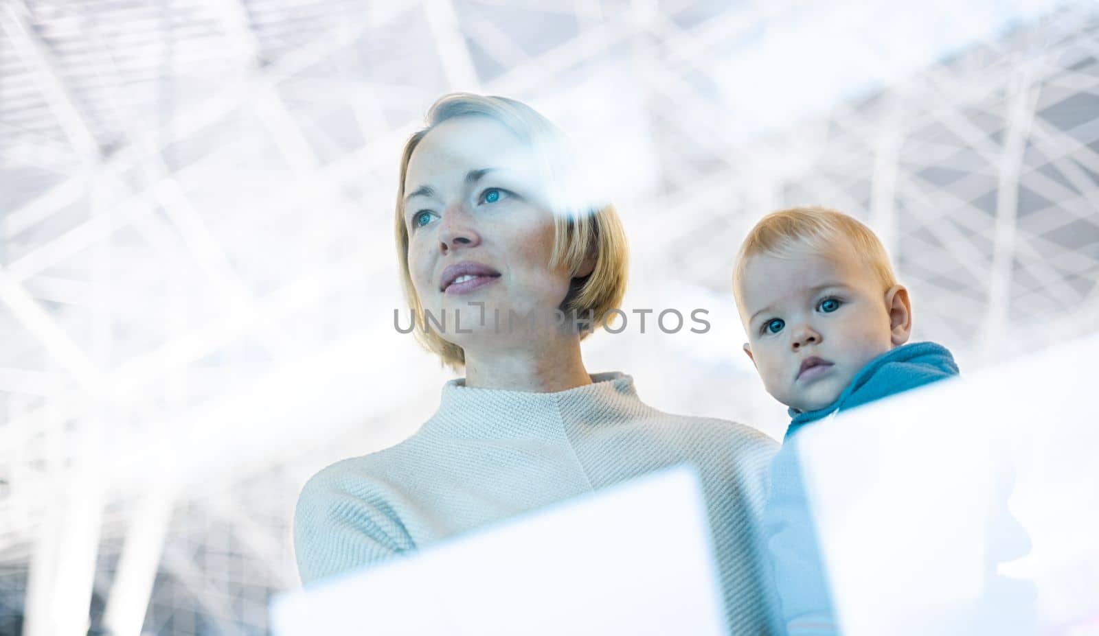 Thoughtful young mother looking trough window holding his infant baby boy child while waiting to board an airplane at airport terminal departure gates. Travel with baby concept