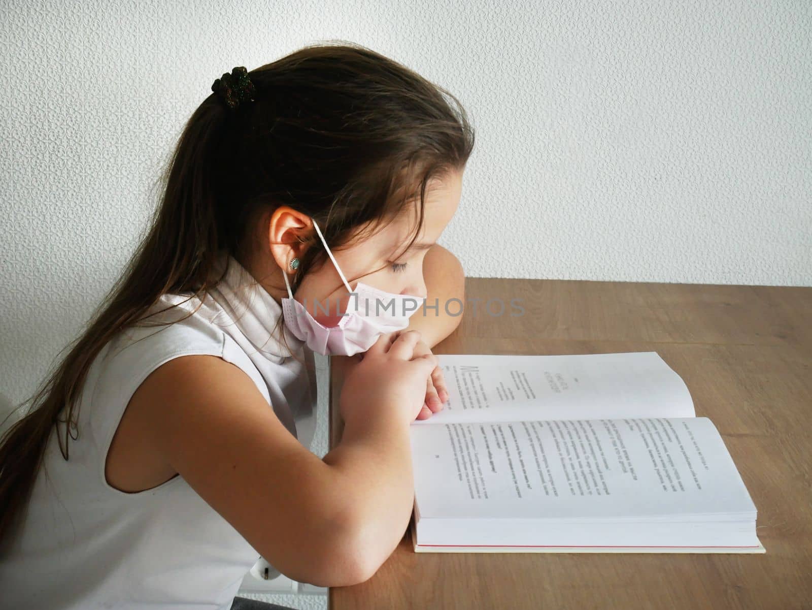 A girl reads a book at home with a medical mask on her face and doing school homework during the Covid-19 pandemic. a protective mask on the girl's face from the corona virus by Jyliana