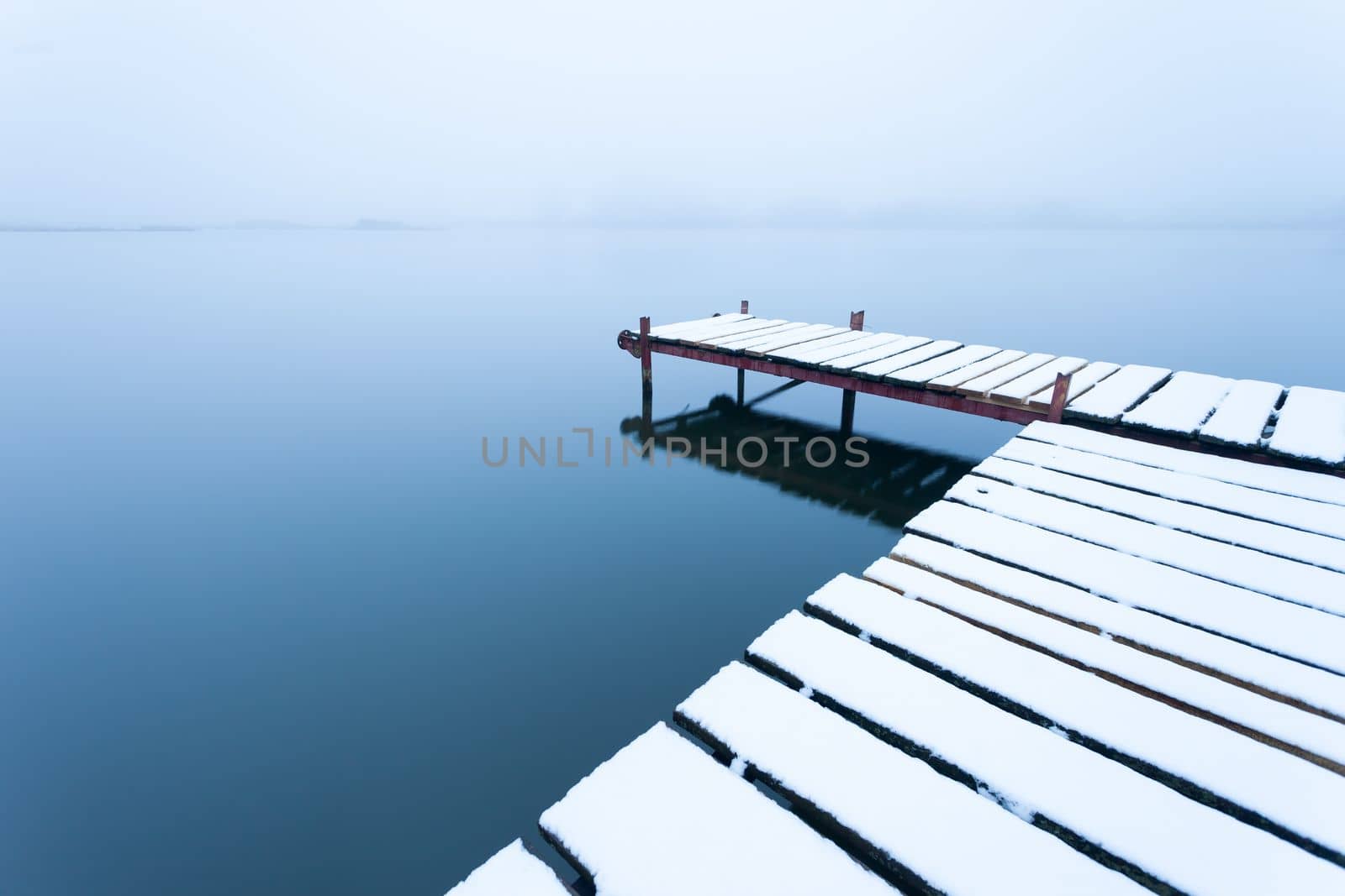 A snow-covered pier with a calm lake on a foggy day by darekb22