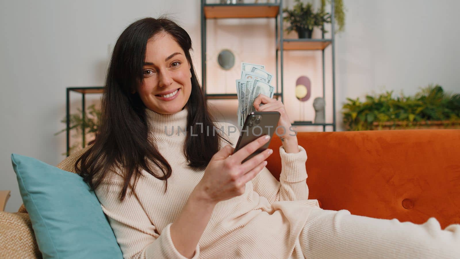 Planning family budget. Young happy brunette woman counting money dollar cash use smartphone calculate domestic bills at home. Joyful girl satisfied of income saves money for planned vacation gifts