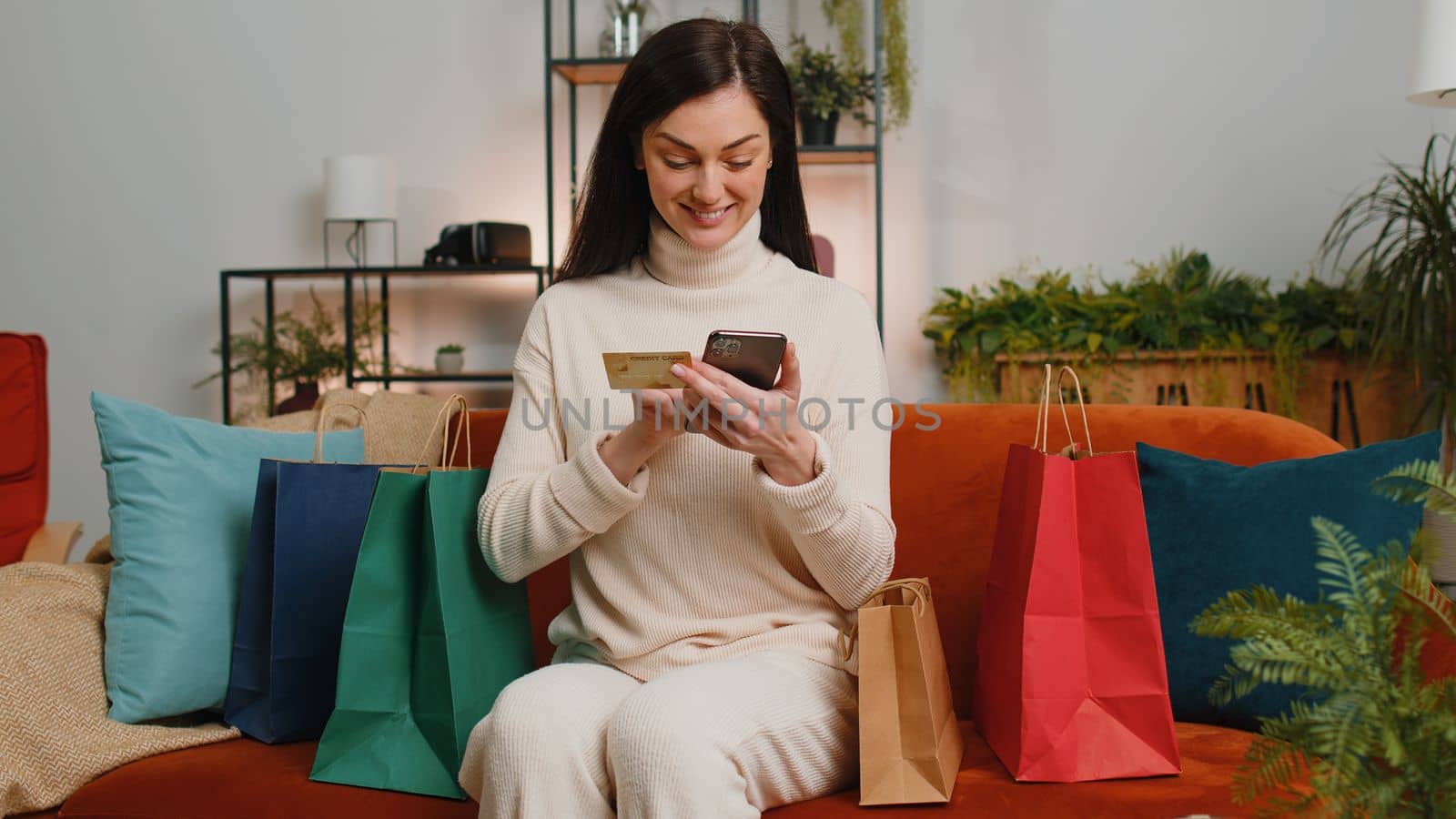 Happy shopaholic consumer young woman sitting with shopping bags at home apartment sitting on couch making online order, payment with credit plastic bank card. Discount sale on internet store mall