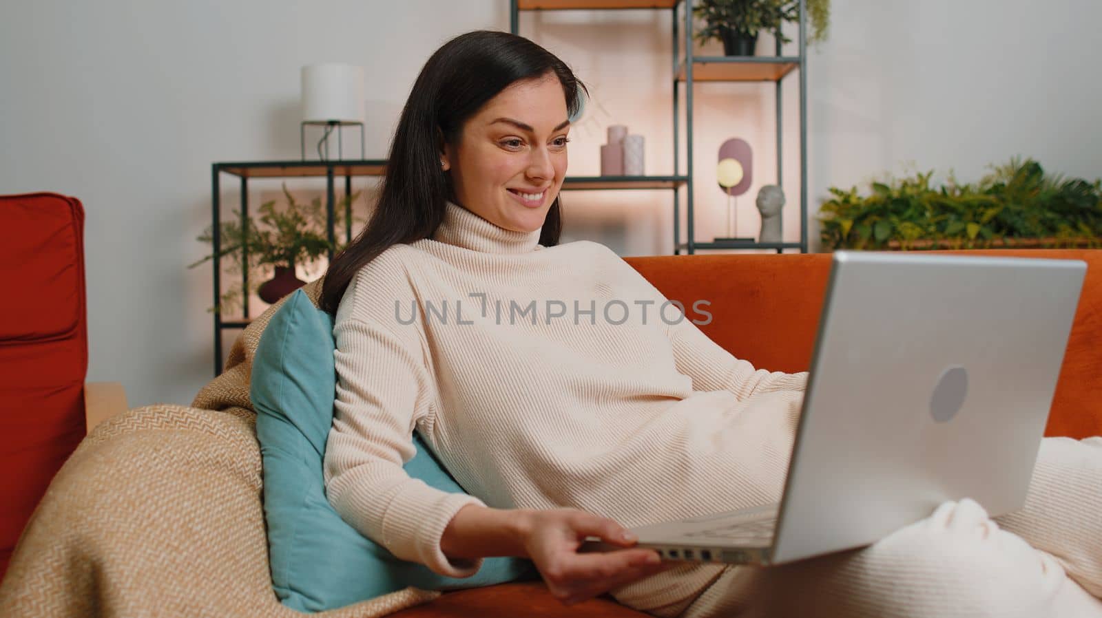 Young brunette woman sitting on couch looking at camera, making video webcam conference call with friends or family, enjoying pleasant conversation. Girl laughing waving hello at home living room