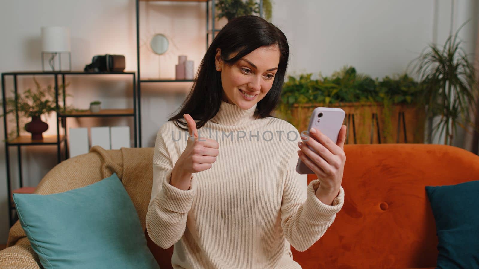 Young woman making smartphone call online video webcam conversation with friends sitting on couch at home in living room apartment. Happy excited girl enjoying mobile loudspeaker talking indoors