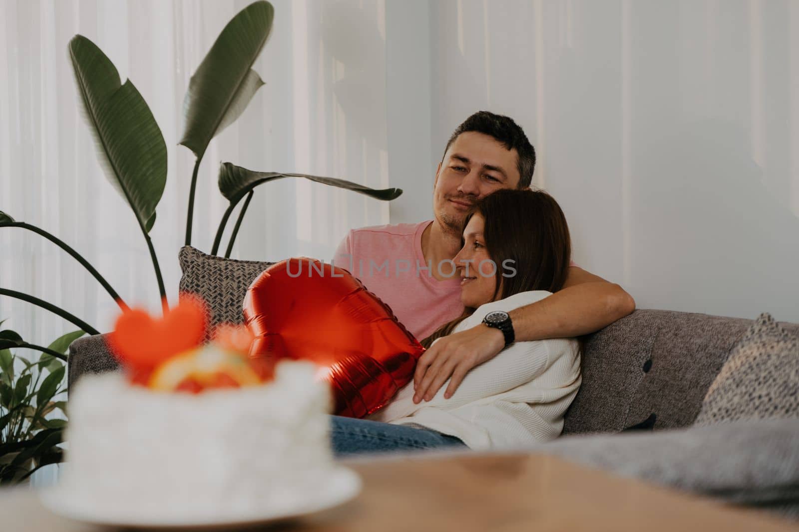 Beautiful young couple sitting on a couch, hugging on romantic date on Valentine day at home. Wife and husband celebrate with heart shape ballon, cake, flowers by Ostanina