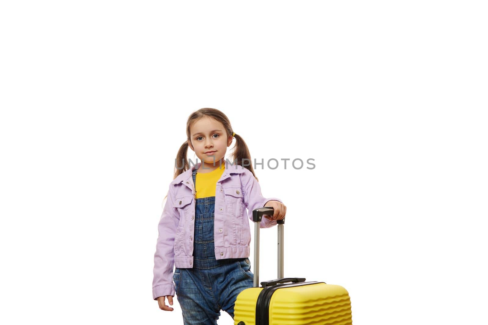 Caucasian cute baby girl, little traveler child dressed in casual denim overalls and purle jacket, standing near her yellow suitcase isolated over white background. Tourism. Travel. Advertising space