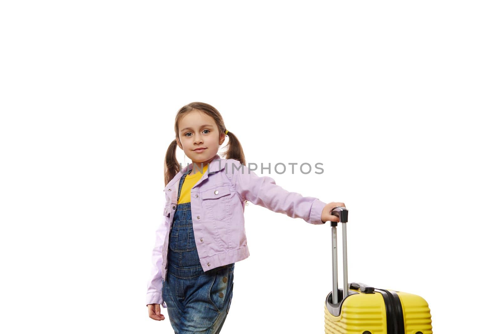 Caucasian happy little child girl with two ponytails, in purple denim jacket, looks at camera, posing with yellow suitcase over white background with copy space for ad. Tourism. Kids travelling alone