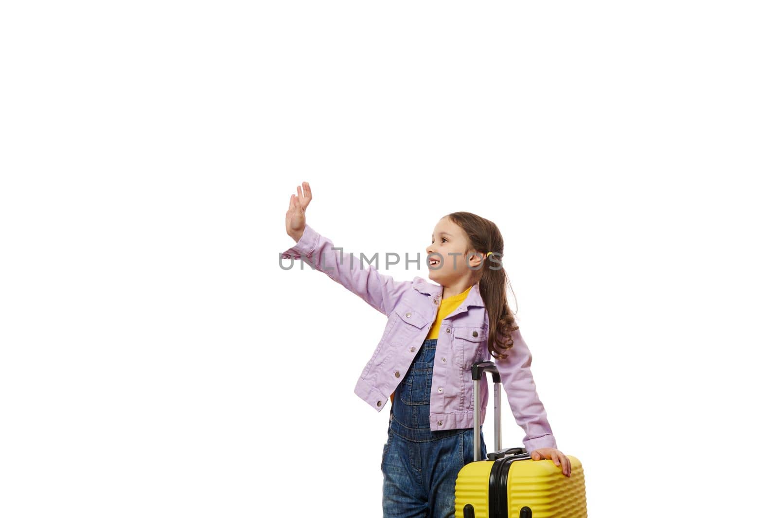 Cheerful adorable little child girl, traveler in purple jacket and blue denim overalls, with yellow suitcase, waving hello with hand, looking at copy space on white. Summer vacation and travel concept