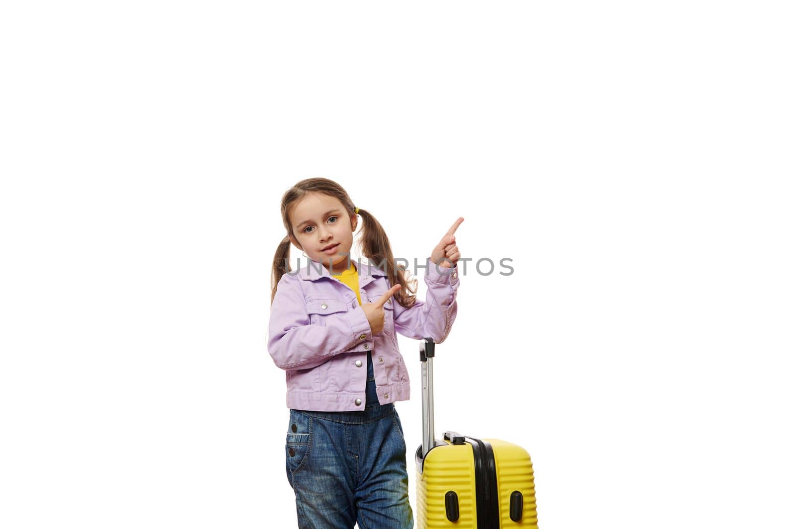 Little child girl in blue denim overalls and purple jacket, standing near her yellow suitcase, looking at camera and pointing forefingers at copy space for advertising text, on white background