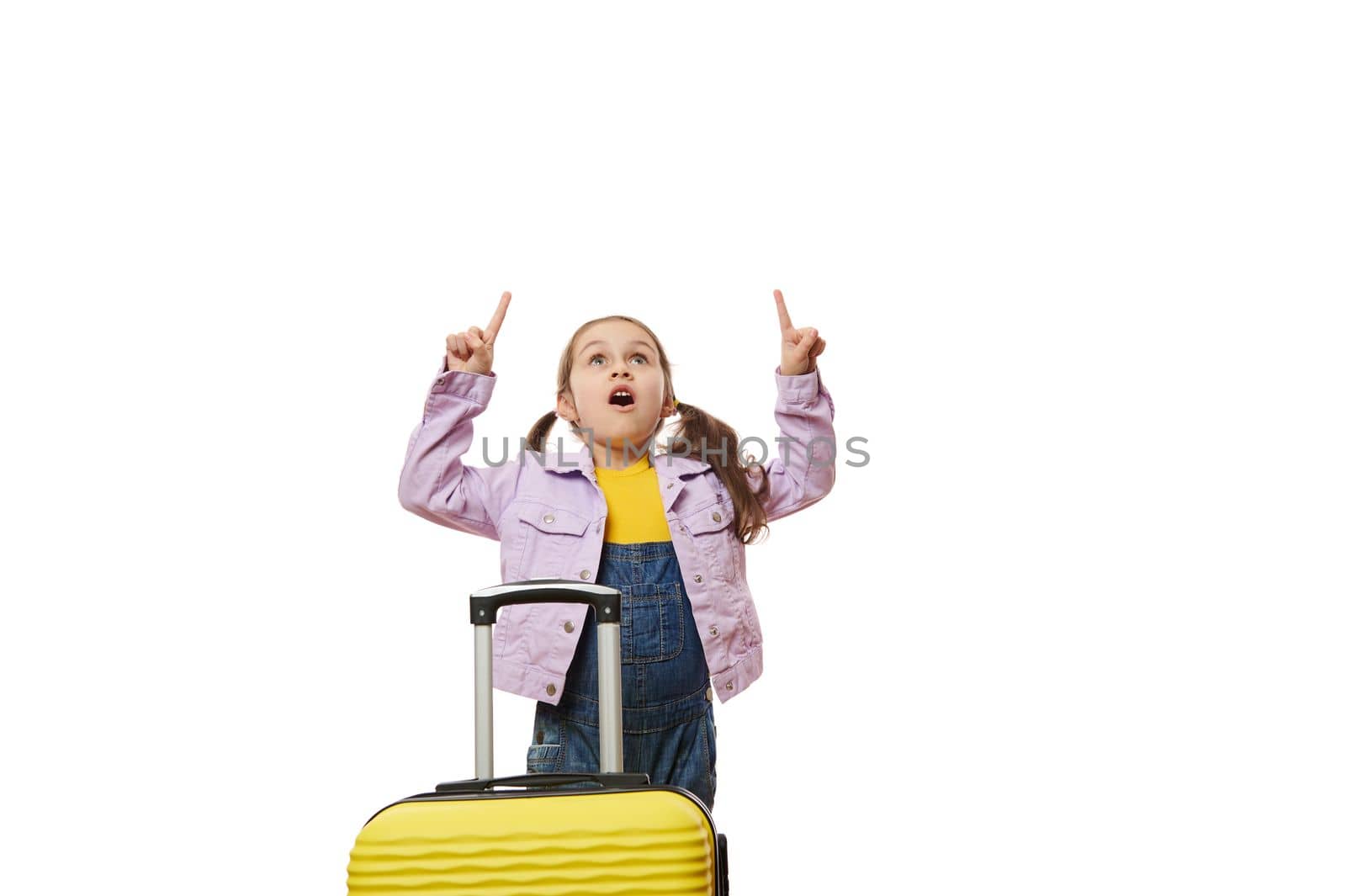Amazed little traveler girl, dressed in purple jacket and blue denim overalls, standing behind yellow suitcase, expressing wow emotion looking up, pointing forefingers at copy ad space, white backdrop