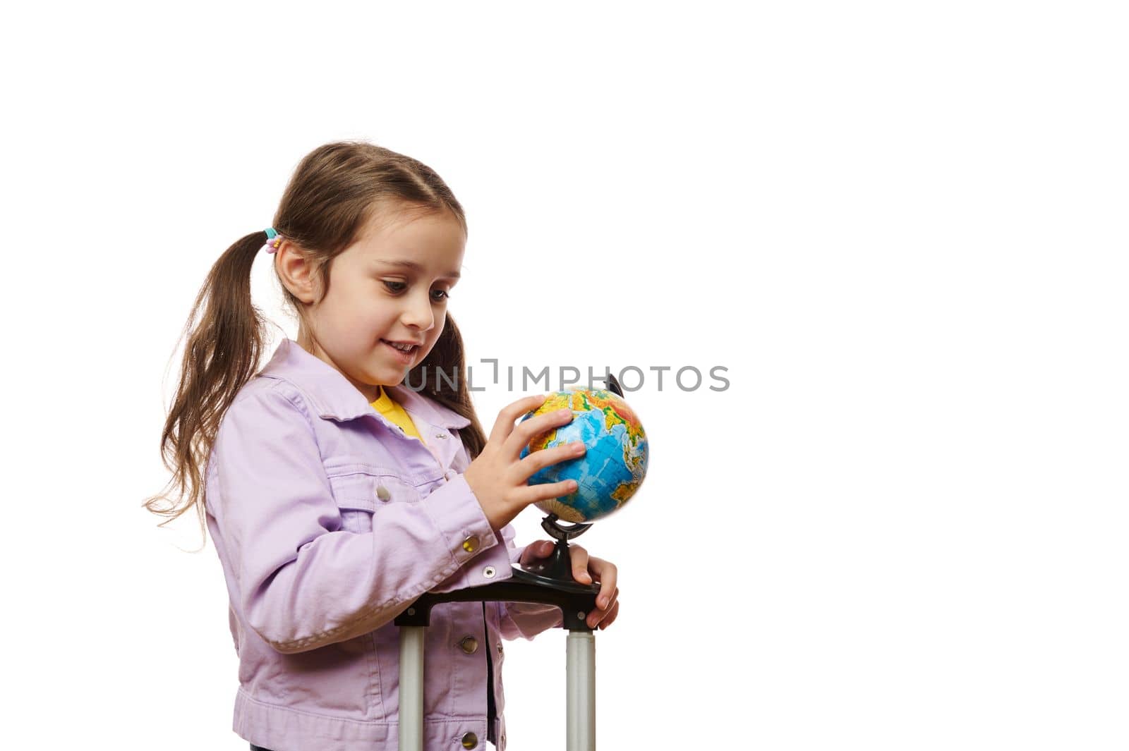 Happy European kid girl 5-6 years old with a suitcase, pointing with her finger at the globe, isolated on white background. Little child passenger traveling abroad for the weekend. Air travel concept