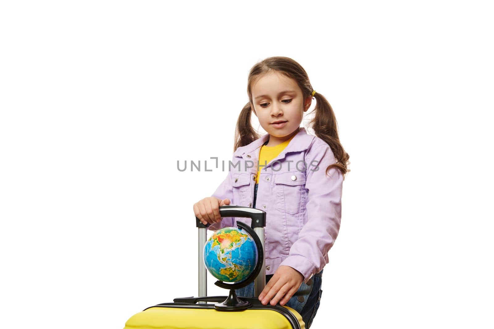 Beautiful Caucasian little child traveler tourist, lovely baby girl with two ponytails, in purple denim jacket, posing with globe and yellow suitcase on white background. Worldwide tourism concept