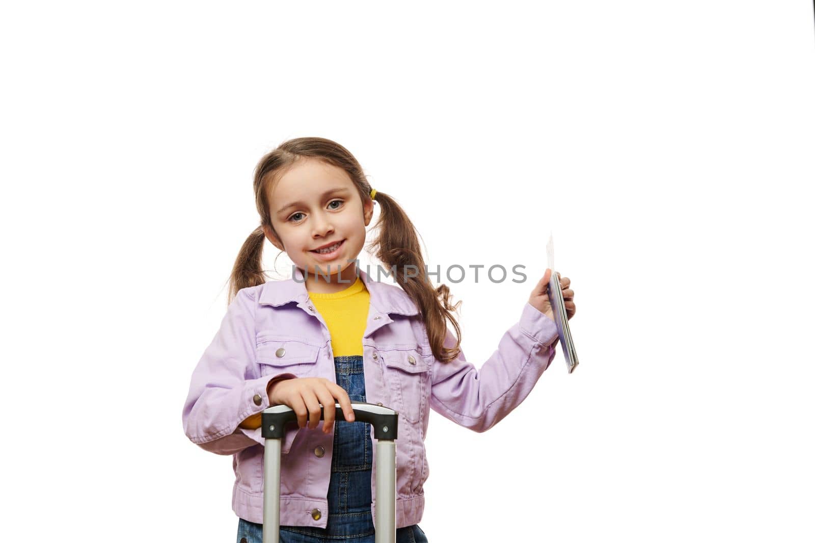 Adorable little traveler girl in purple jacket, holding suitcase and boarding pass, smiles looking at camera, on white by artgf